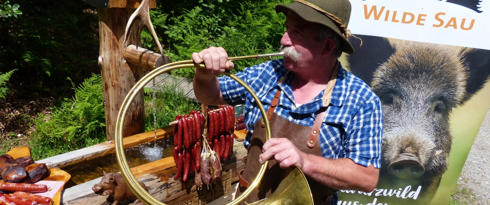 Mann spielt Jagdhorn. Trägt Filzhut, kariertes Hemd und Lederschürze. Wildschweinsalami hängt an Gestell. Projektbanner mit Wildschwein im Hintergrund.
