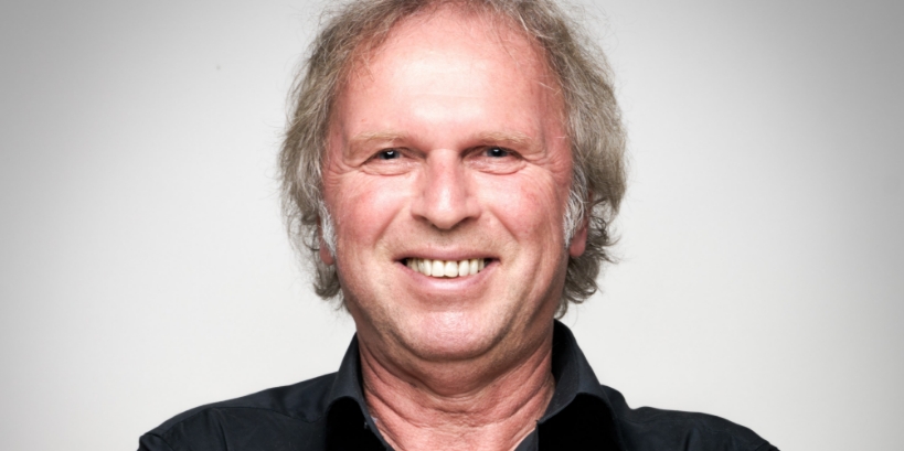 Ein lächelnder Mann mit grauem Haar und schwarzem Hemd vor grauem Hintergrund.