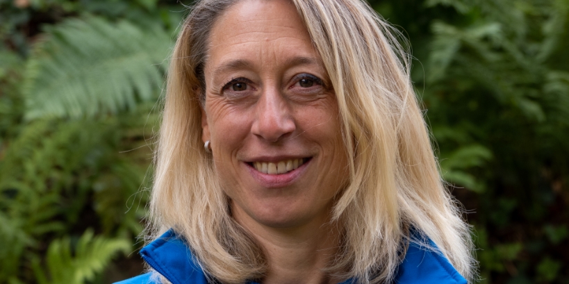Frau mittleren Alters mit schulterlangem, hellem Haar und blauer Outdoor-Kleidung, steht vor grünen Farnen im Hintergrund.