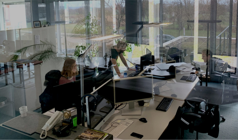 Büroszene mit drei Menschen an einem großen Tisch mit Computern, Fenster bieten Blick nach draußen.