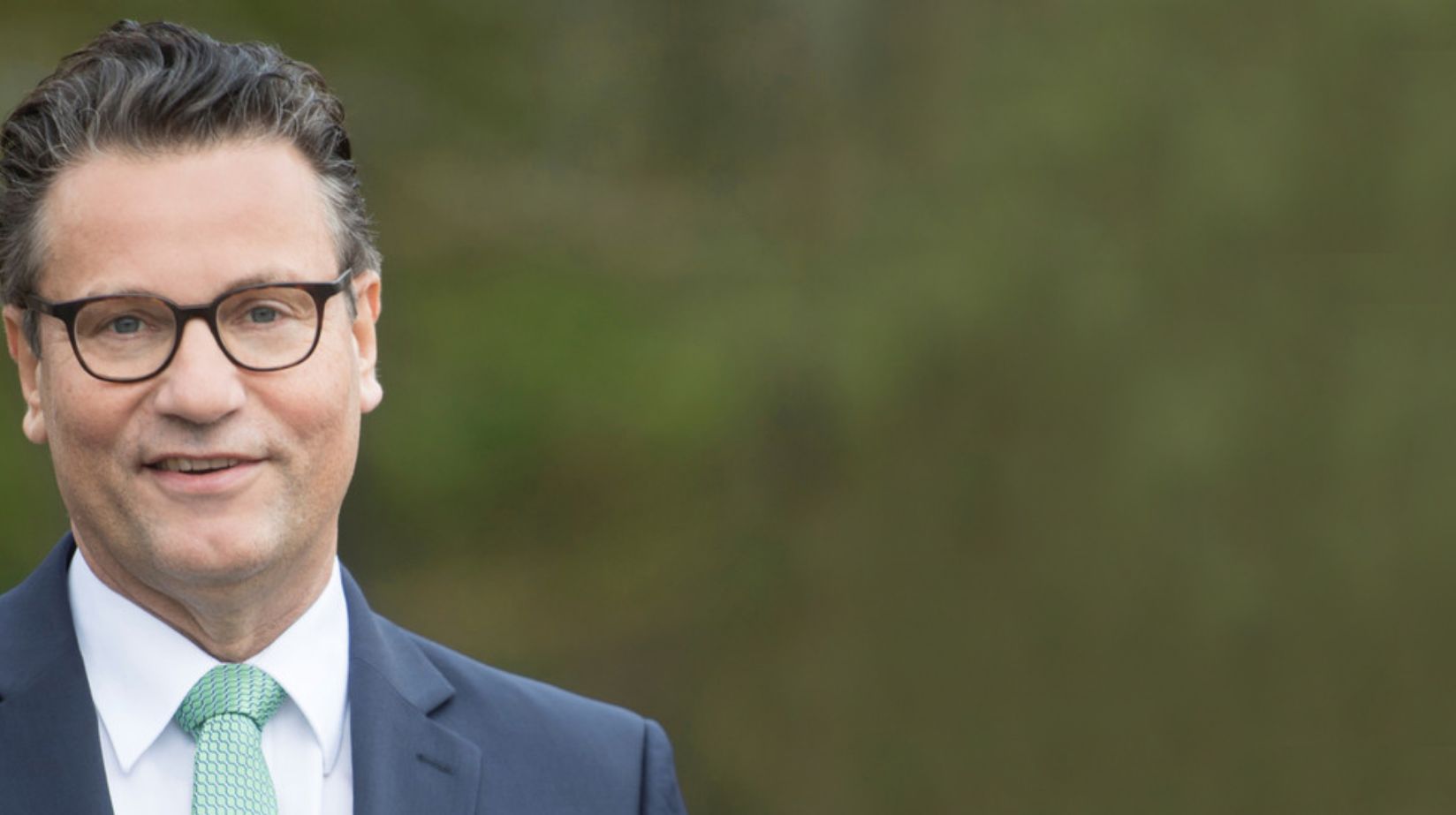 Mann mit Brille, Anzug und grüner Krawatte vor verschwommenem grünen Hintergrund.