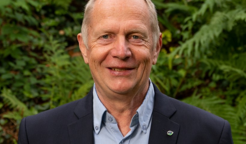 Älterer Mann mit Anzugjacke und Hemd vor grünem Laubhintergrund, lächelt freundlich direkt in die Kamera.