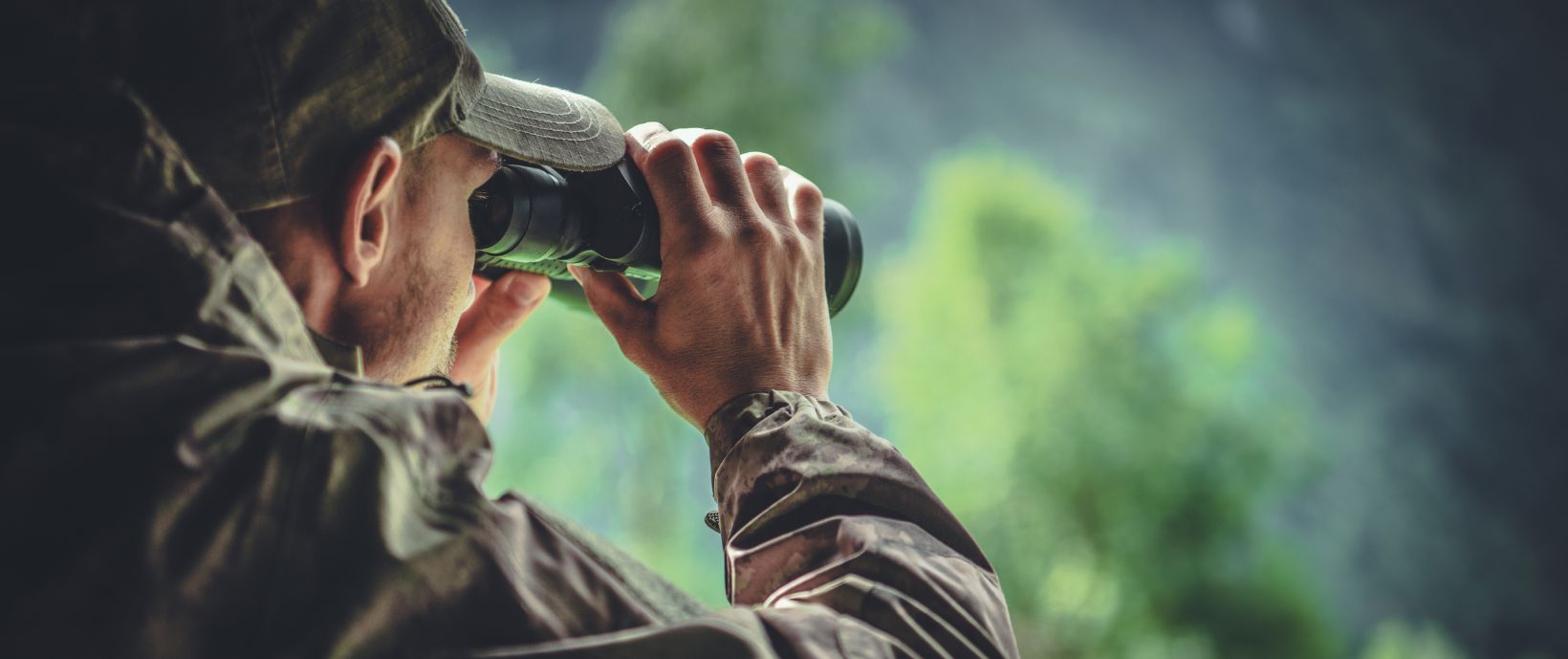 Ein Mann in olivfarbener Outdoor-Kleidung beobachtet mit einem Fernglas die Umgebung, im Hintergrund unscharfe Bäume.
