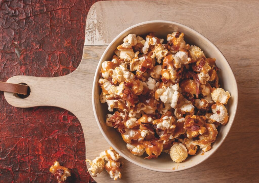 Eine Schale mit karamellisiertem Popcorn und Speck auf einem hölzernen Schneidebrett mit rotem Muster.