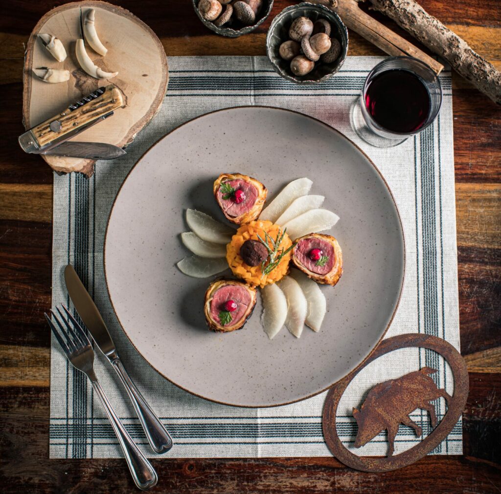 Runder Teller mit Wildschweinrouladen, garniert mit rosa Zwiebeln und Dill, Birnenscheiben, Glas Rotwein. Tischset aus Holz und kariertes Platzdeckchen.