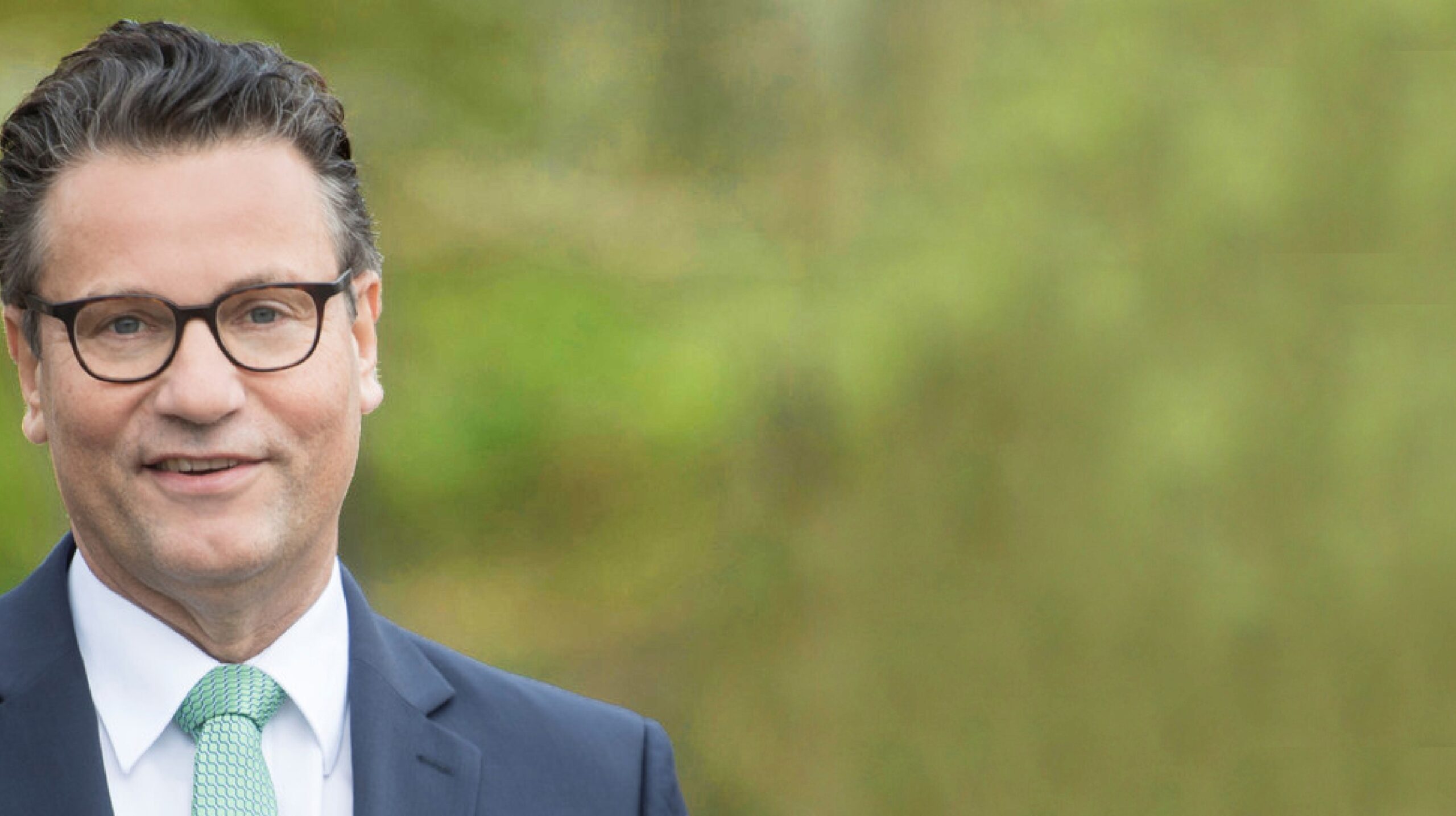 Mann im Anzug mit weißem Hemd und grüner Krawatte vor unscharfem grünen Hintergrund, trägt Brille und leichte Wellen im Haar.
