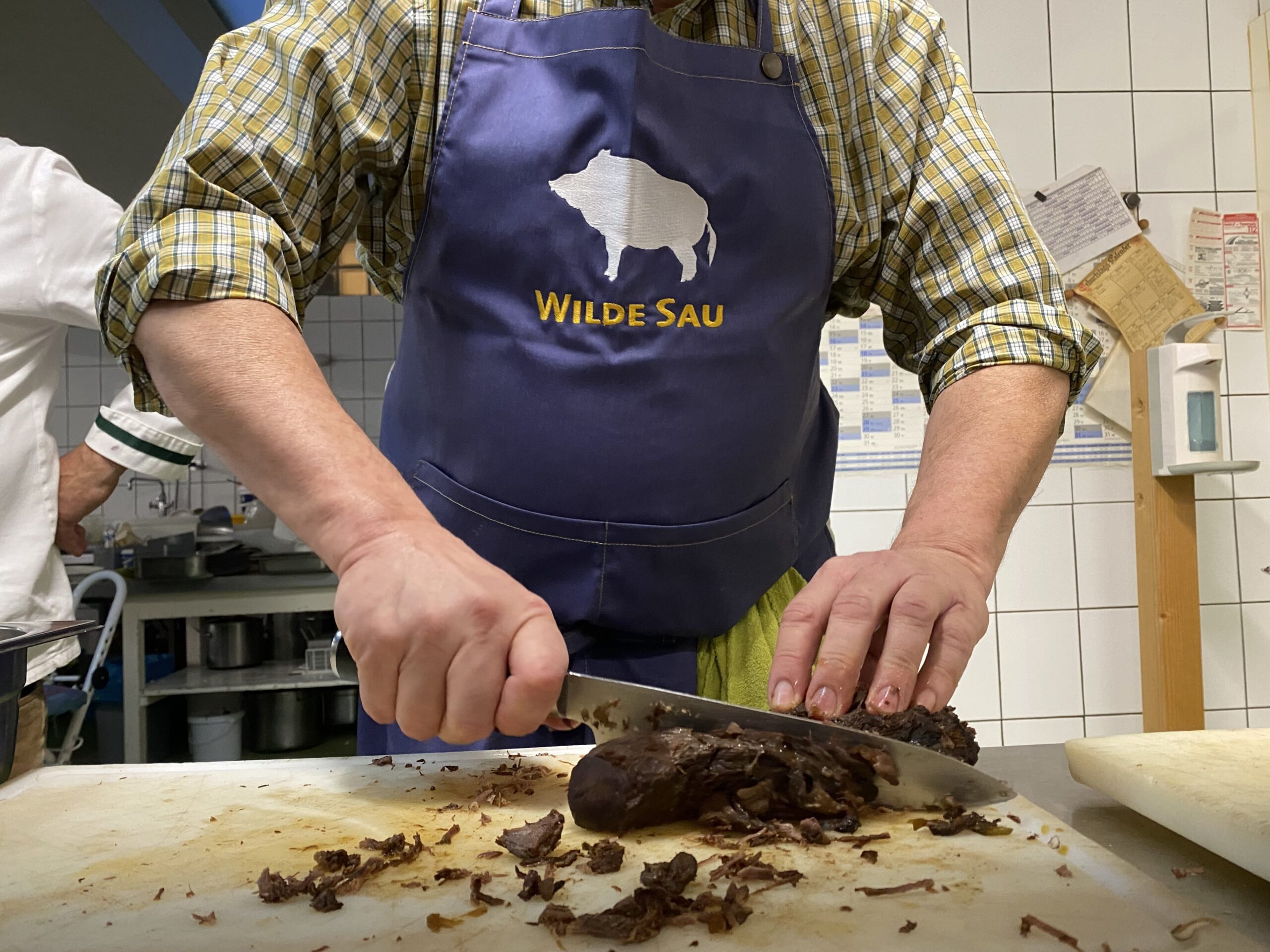 Person mit blauer 'Wilde Sau'-Schürze schneidet Wildfleisch in einer gefliesten Küche.