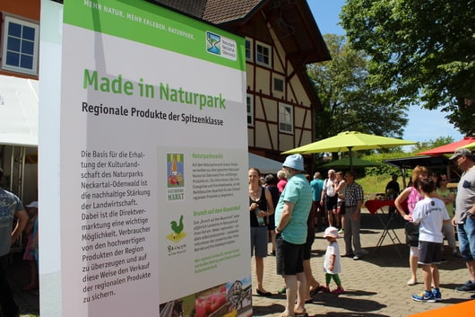 Naturparkmarkt mit Plakat „Made in Naturpark“, Menschen erkunden den Markt, Fachwerkhaus im Hintergrund.