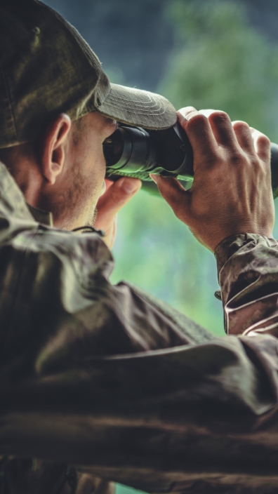 Mann in jagdlicher Kleidung mit Fernglas in der Natur, Bäume im Hintergrund.