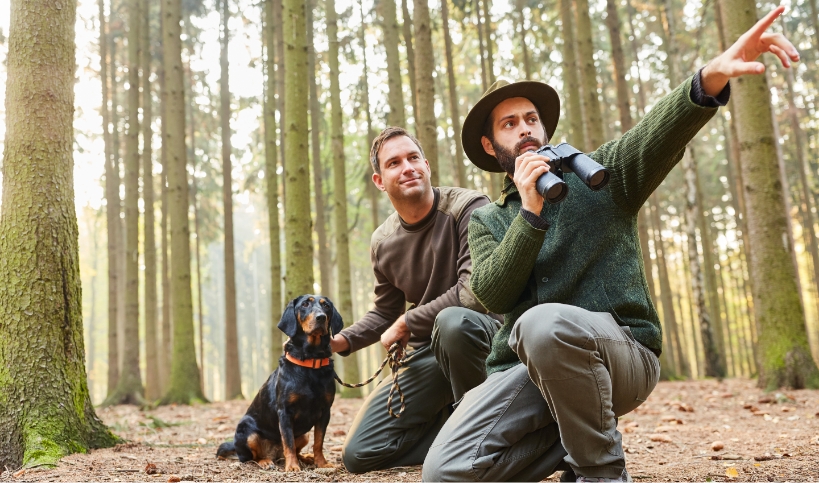 Zwei Männer mit Fernglas und Hund in einem Nadelwald, kniend auf dem Waldboden, einer zeigt in die Ferne.