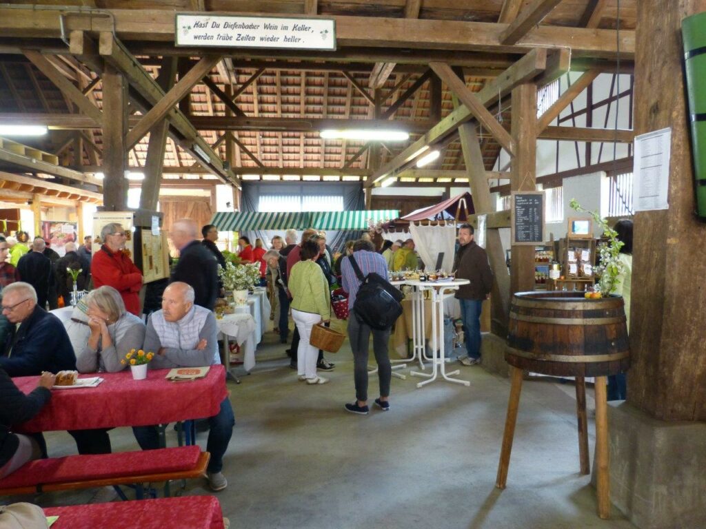 Besucher erkunden Marktstände in der rustikalen GenussScheune, Holzbalken und rote Tische sichtbar.