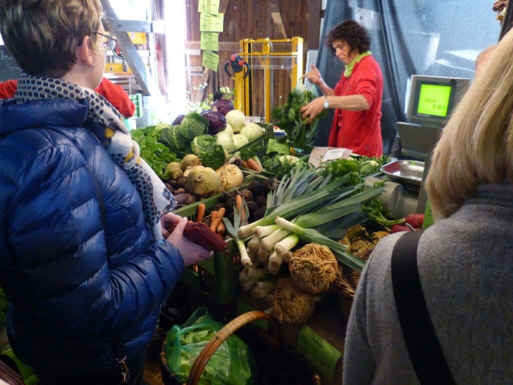Verkäuferin an einem Marktstand mit Lauch, Sellerie, Kohl und Karotten, Kunden im Vordergrund.