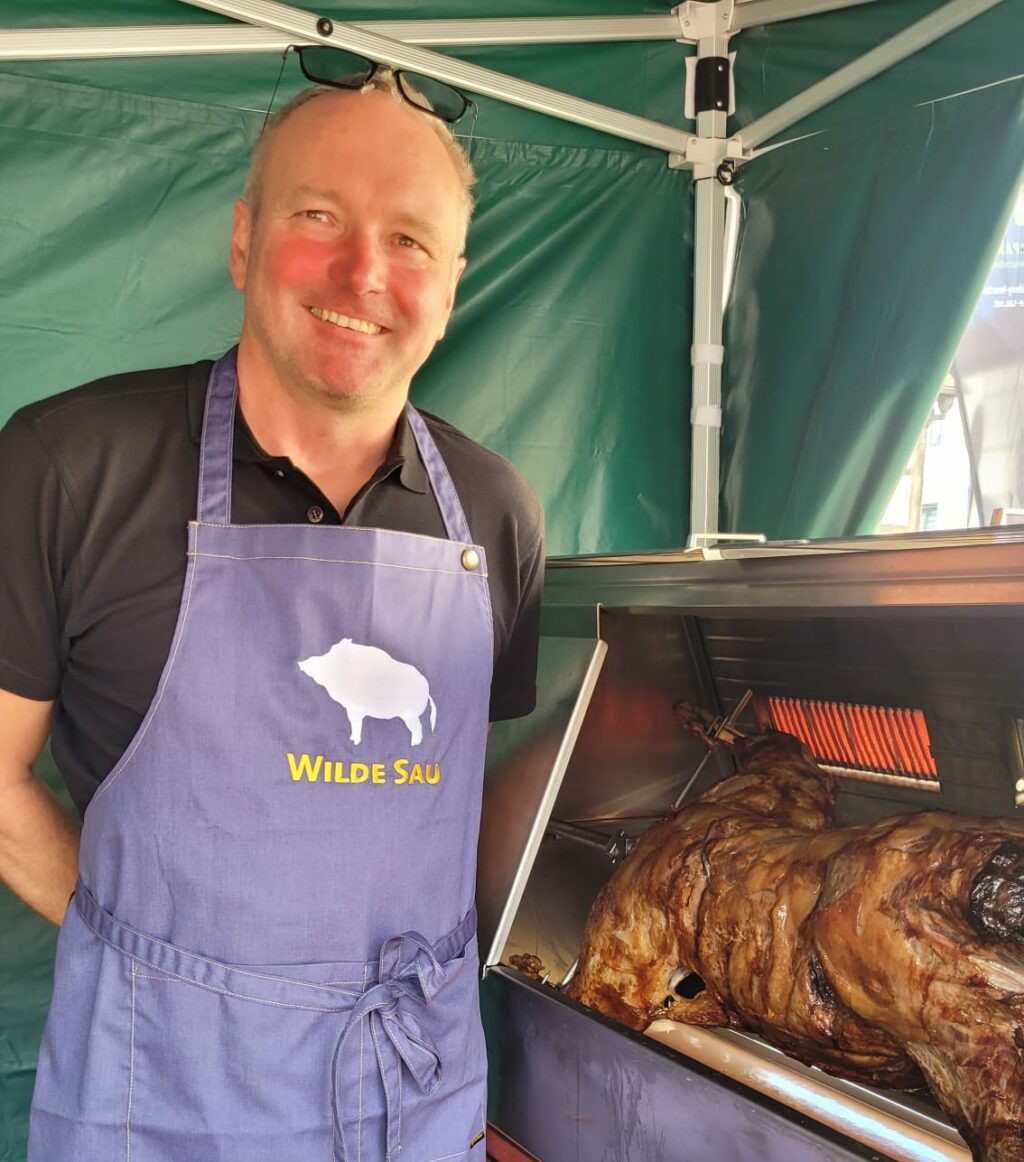 Grillmeister mit 'Wilde Sau'-Schürze am Grill mit Wildschweinfleisch, lächelt.