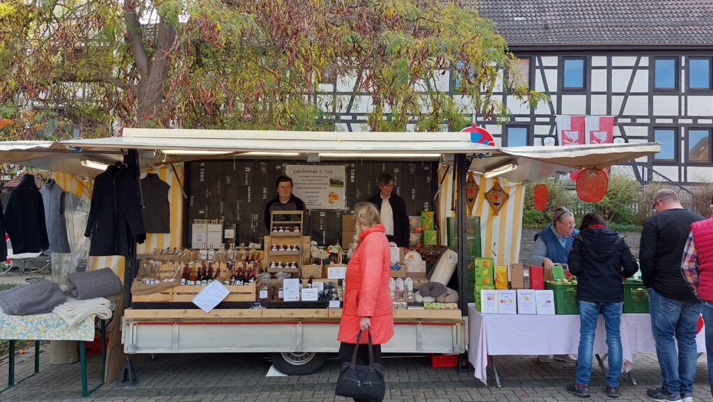 Marktstand auf Event mit Flaschen, Gläsern und Besuchern, Fachwerkhaus im Hintergrund.