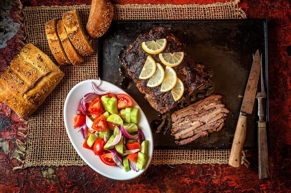 Gyrosschichtbraten mit Zitronen, frischem Salat und Brot auf rustikalem Hintergrund.