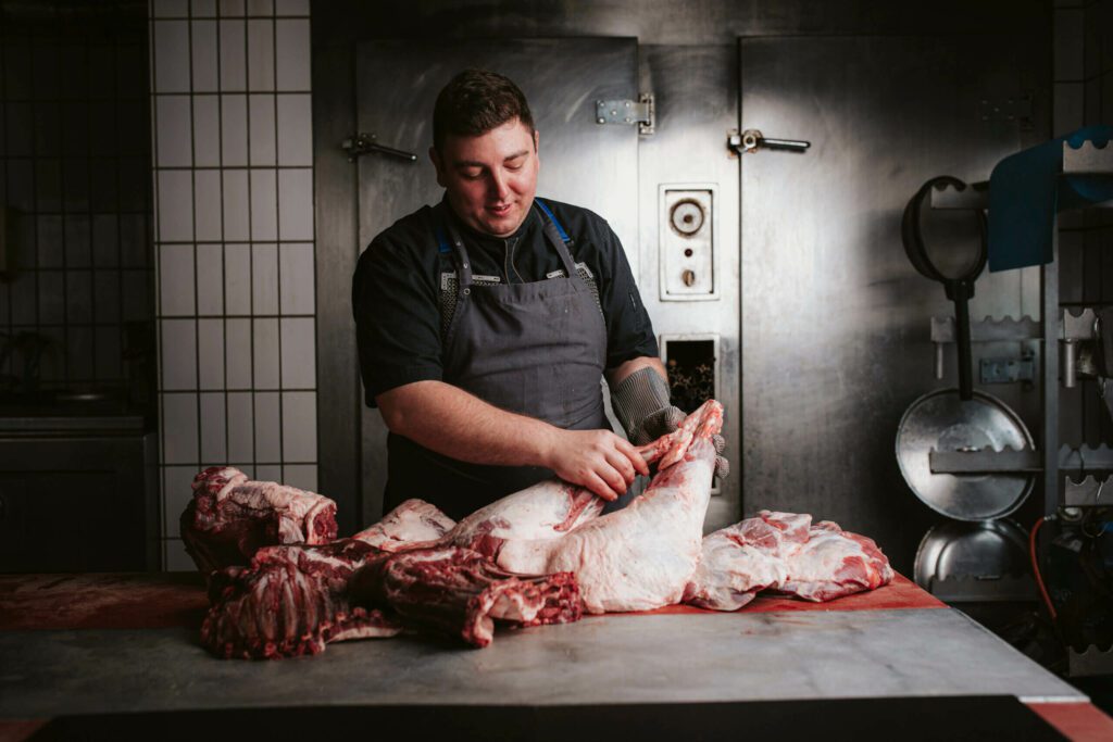 Ein Metzger, der ein großes Stück Wildschweinfleisch auf einem Tisch bearbeitet. Er trägt Schürze und Schnittschutzhandschuh.