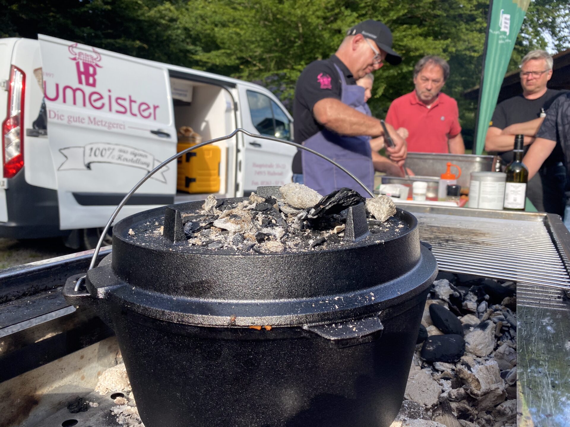 Gusseiserner Dutch Oven mit Kohle auf Grill, Hintergrund mit Männern und Lieferwagen 'Umeister'.