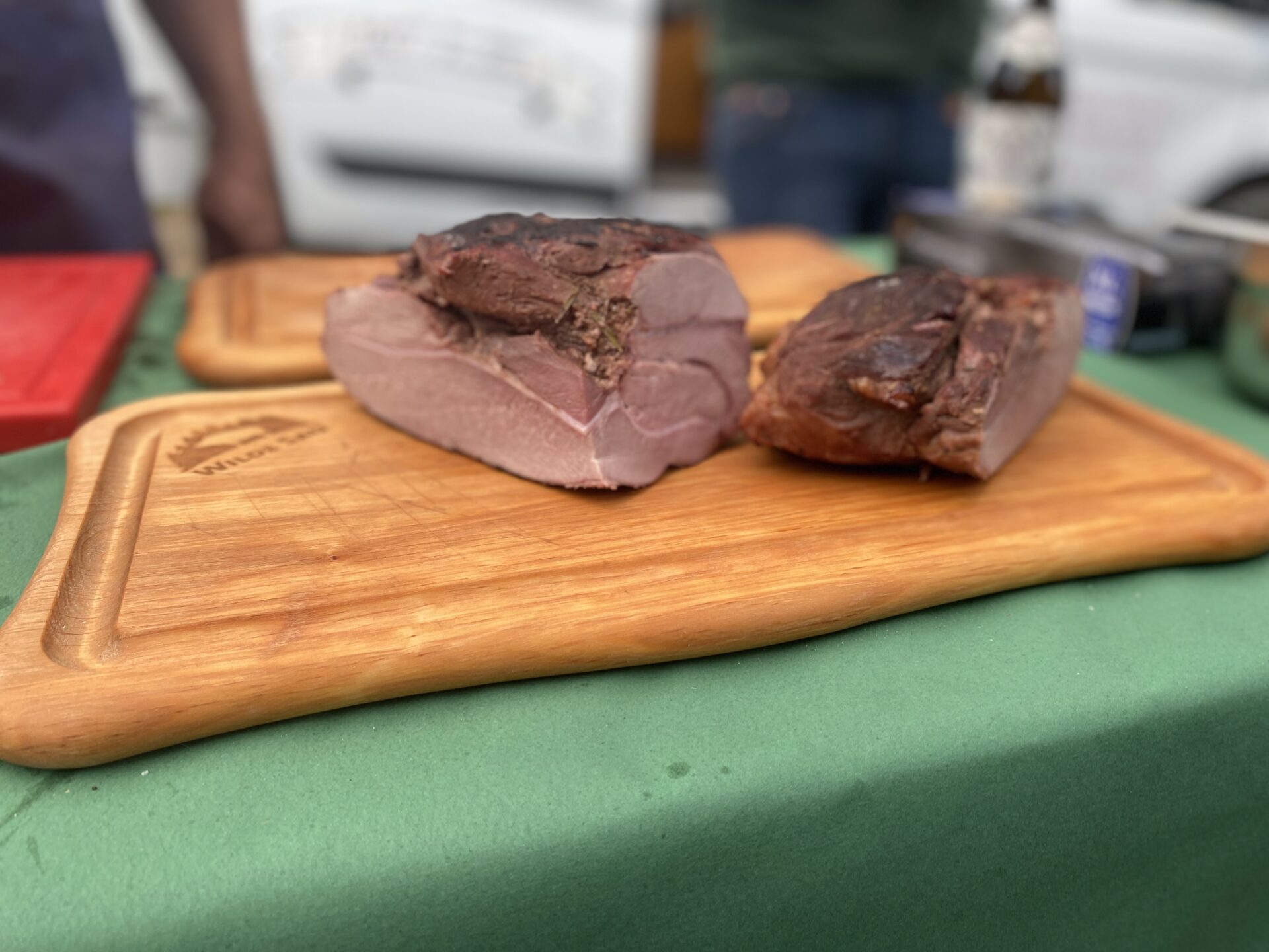 Zwei große Stücke Wildschweinbraten auf einem Holzbrett auf grünem Tisch, Marktatmosphäre im Hintergrund.