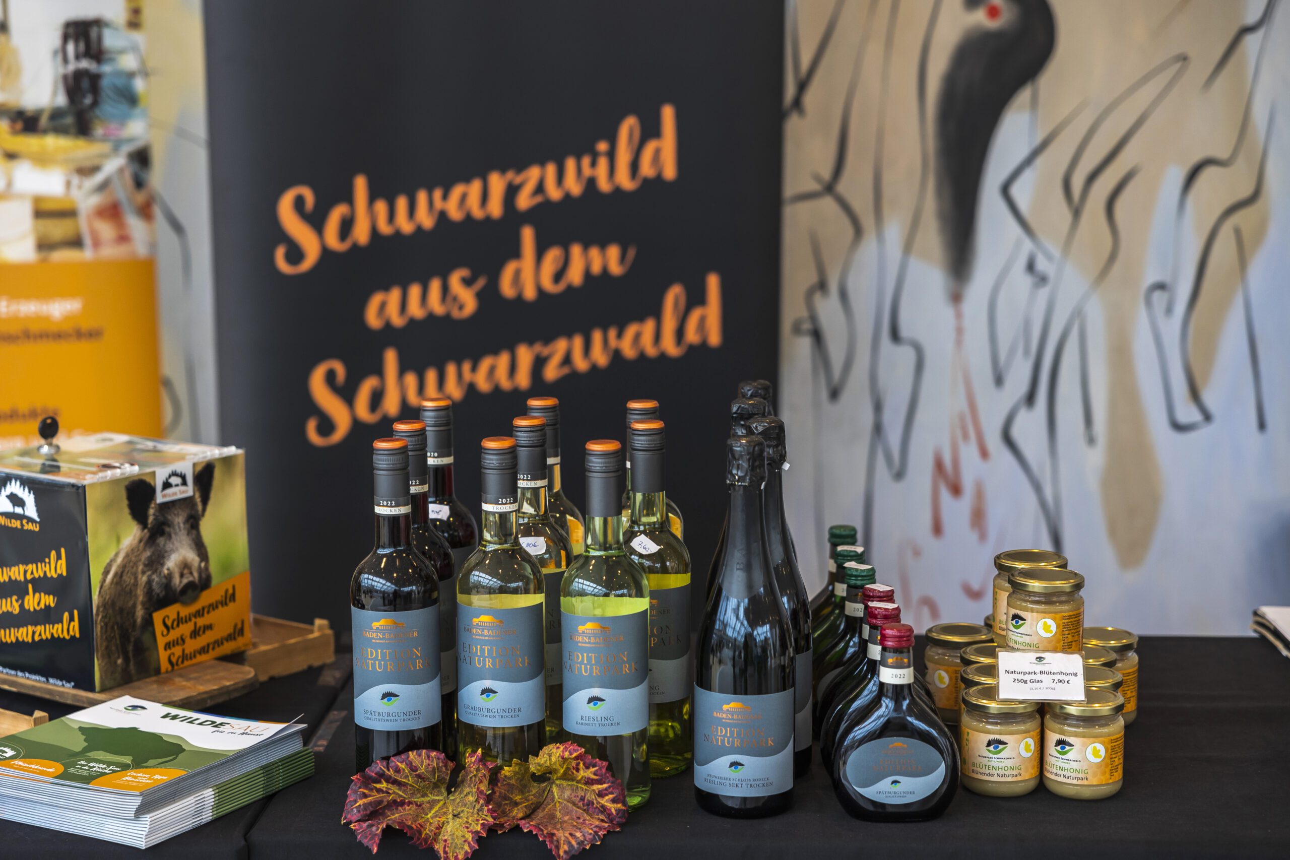 Produkte auf einem Tisch einer Messe: Flaschen Wein, Sekt, Konfitüren und Liköre, Banner mit 'Schwarzwild aus dem Schwarzwald'.